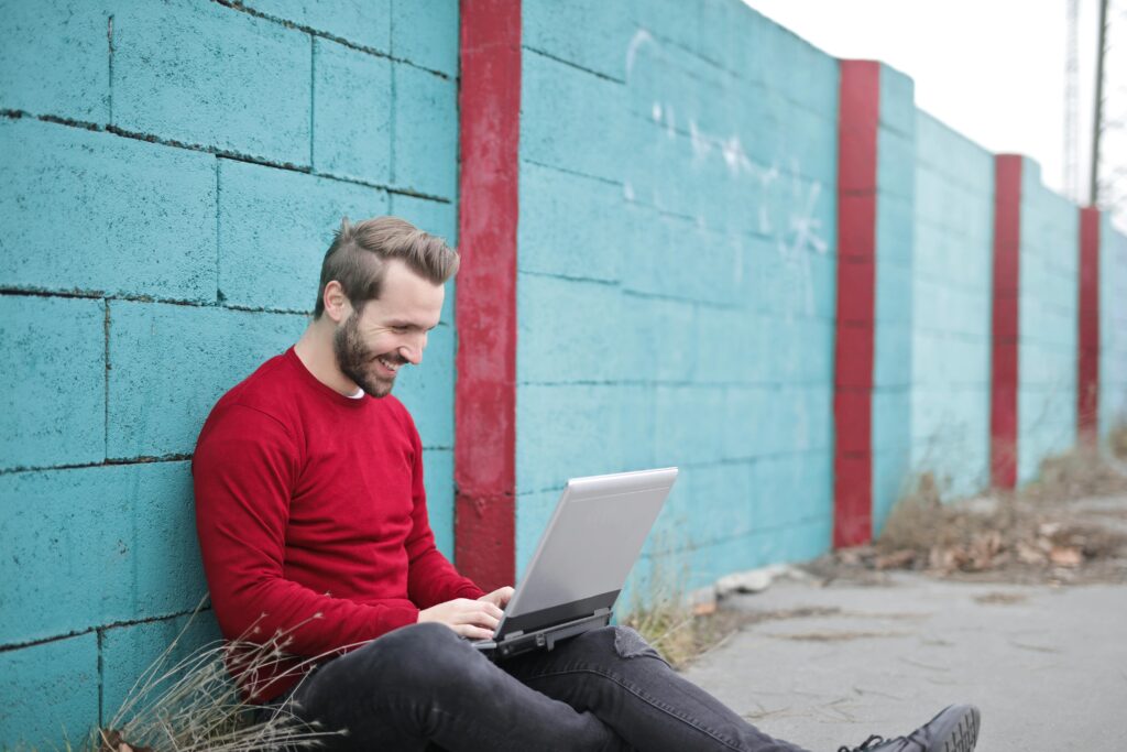 A man smiling and using a laptop outdoors against a vibrant blue wall, representing remote work.